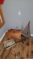 Photo showing the vintage brown woven leather handbag, model sailboat with striped sails, and two metal hooks on a wooden floor near a wall.
