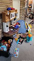 Full view of the Little Tikes play kitchen stove with black bin of kitchen toys and other toys around