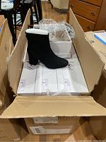 Open large cardboard box containing multiple white shoe boxes with black faux suede ankle boots displayed on top, showing the boot shape and heel.