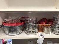 Shelf with various sizes of clear glass food storage containers, some with red lids stacked on top or next to them, plus small clear bowls and lids in an organizer basket.