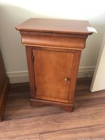 Front view of small wooden cabinet showing molded top and base trim, single closed door with metal knob.