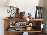 Wide view of decor lot items on wooden shelves and top surface including metal lamp, wooden key hanger, pottery jug, oil lamps, wall plaques, mosaic plates, coaster set, small clock, cast iron door stopper and chair legs.