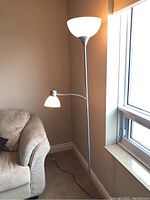 Full view of the 73-inch floor lamp standing beside a couch next to a window, showing the upper torchiere light on and the gooseneck reading lamp below it also lit.