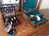 Overview of jewellery boxes, watches arranged inside and outside cases, rosaries and assorted earrings on dresser top.