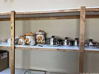 Shelf view showing three ceramic toadstool canisters, three stainless steel pots with lids, one stainless steel pan with lid, and two white serving dishes