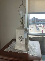 Full view of the 29 inch tall ivory ceramic table lamp on a lace doily, showing its square decorative cut-out design and bulb socket with harp.