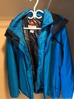 Photo showing light blue Columbia insulated jacket hanging on a white hanger with a zip closure and hood.