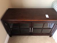 Front view of dark wood television A/V cabinet showing glass double doors with metal handles and overall shape.