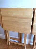 Four wood TV trays stacked and standing upright against a wall showing light wood finish and folded legs.