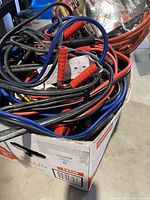 Photo showing multiple booster cables with red and black clamps and blue and black cables stored in a box.