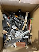 Top-down view inside cardboard box full of various kitchen cutting knives and utensils with mixed handles.