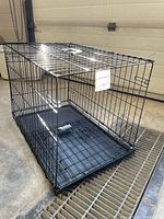 Side perspective of a black metal wire dog cage with a removable plastic tray base, folded flat bottom and a metal latch door on the side.