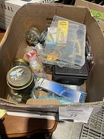 Box containing fishing gear, tackle boxes, jars with fishing hooks, fishing lures in packaging, and spools of fishing line.