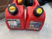 Two red plastic 5L gas jugs side by side on concrete floor showing front labels and caps.
