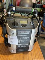 Front and top view of Motomaster Eliminator booster pack with air compressor, showing handle and control panel with LCD screen, switch, and USB ports.