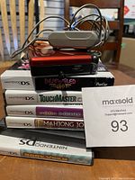 DS games stacked with red DS Lite console base and power adapter on top, showing game titles on cases and charger cable coiled