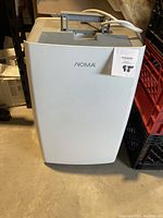 Front view of the white Noma dehumidifier in a storage setting on the floor showing its compact rectangular design with handle and power cord placed on top.