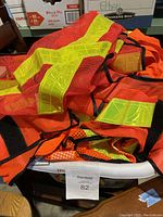 Image showing a white basket filled with multiple fluorescent orange mesh safety vests with reflective yellow stripes and black trim, placed on a chair and surrounded by boxes.