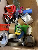 Box view showing multiple rolls of tape in red, brown, green, blue, white, and silver, with several pairs of scissors inside.