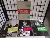 Front view of the three Andy Warhol auction catalogues with the original slipcase behind them, showing the covers featuring black and white photos of Warhol with colorful text bands.