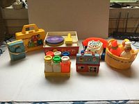 Full view of seven vintage Fisher-Price toys including plastic carrying case, record player, rotary phone, salt and pepper shakers, bath toy, cash register, and washing machine toy.