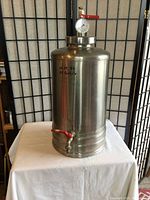 Full view of the large cylindrical stainless steel container with pressure gauge on top and spigot tap at bottom, placed on a white cloth-covered table.