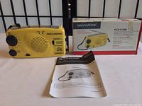 Yellow Magnasonic PCR110M AM/FM radio with solar panel and hand crank, shown with user guide and original box in background.