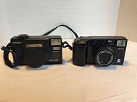 Two Minolta film cameras side by side with wrist strap visible, plastic body, compact style