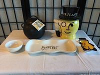 Full view of all Mr. Peanut ceramic collection items on a white cloth: yellow cookie jar, black bean pot, cream tray, small bowl, and peanut-shaped dishes.