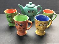 Photo of green teapot with smiling cartoon face and four colorful cartoon face pedestal mugs arranged around it on black surface.