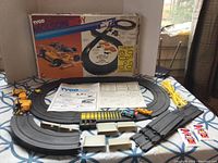 Full view showing assembled slot car racing track with two slot cars, yellow supports, guardrails, connectors, original box, and instruction sheets on a table.