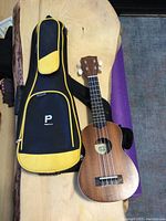 Full view of the small ukulele with natural wood finish next to black and yellow padded soft carrying case