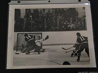 Black and white vintage hockey photo showing a player shooting on goal with goalie and a defenseman near the net. Visible crowd behind glass in stands.