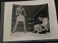 Front side of a black and white sport press photo showing Sugar Ray Robinson and Rocky Graziano in a boxing match, Robinson standing and Graziano down on the canvas.