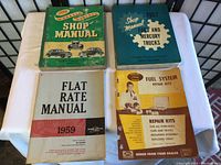 Photo showing the four vintage vehicle maintenance manuals laid out on a white tablecloth, featuring manuals for Ford, Monarch, Mercury vehicles, flat rate manual and fuel system repair kits.