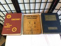 Three vintage vehicle maintenance manuals arranged side-by-side: Chilton's 1959 Auto Repair Manual (red cover), Shop Manual Supplement (worn beige softcover), and Motors Auto Repair Manual (blue hardcover).