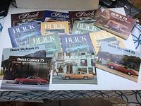 Photo showing a variety of vintage Buick brochures from the 1970s, including Skyhawk, Century, Apollo, LeSabre, Electra, Riviera, and Regal models displayed on a table.