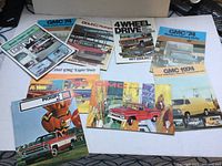 Lot of vintage GMC vehicle salesman brochures, mostly pickups and vans from 1967, 1974 and 1977 models laid out on flat surface.