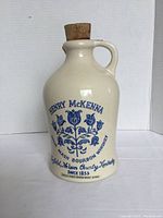 Front view of ceramic finger jug showing Henry McKenna branding and floral design in blue on cream colored jug with cork stopper.