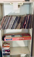 Photo of white shelving unit containing numerous vinyl albums on one shelf, boxed keyboard on shelf below, and VHS tapes stacked on the shelf.