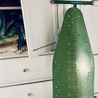 Green metal ironing board leaning against a wall beside the dresser
