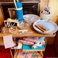 Full view of wooden kitchen cart containing various kitchen items including Thermos, mixing bowl, utensils, plates, place mats, and table cloths.