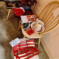 Photo showing three trays in black, red, and rectangular shapes, along with an 18-piece utensil set on and around a wooden chair. Includes ceramic pitcher, bowl, and tea set visible on the chair seat. Additional smaller items like ceramic birds also visible.