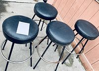 Four round stools with black leather padded seats and black metal legs with chrome footrests photographed outdoors on concrete against a wooden fence.