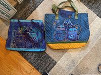 Both Lauren Burch tote bags placed on a carpet. One bag is blue-purple with three cats and purple handles, the other is blue-yellow with green handles and three cats.