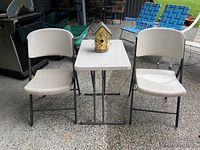 Overall view of folding table with birdhouse on top and two folding chairs on the sides, outdoor setting.
