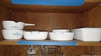 Shelved Corning Ware baking dishes with Blue Cornflower pattern, various sizes and shapes, some with lids and some without, inside a wooden cabinet