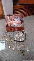 Full view of costume jewelry in and around wooden box showing various necklaces, pins, pendants on carpet.