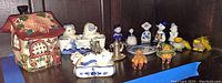 Wide view of shelf containing ceramic house container, Delft blue figurines, small animal figurines, dolls, and plush character faces.