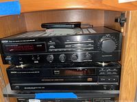 Denon stereo components in a wooden shelving unit, showing receiver and CD player stacked with remotes and cables on top.